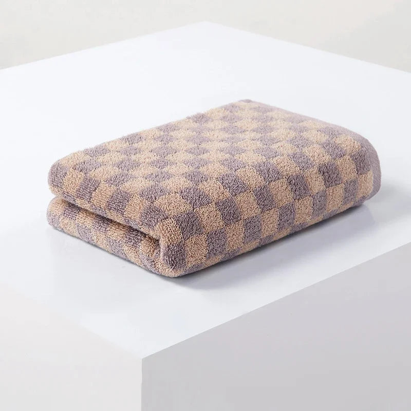 A folded soft plaid cotton bath towel set with a beige and purple checkered pattern on a white surface.