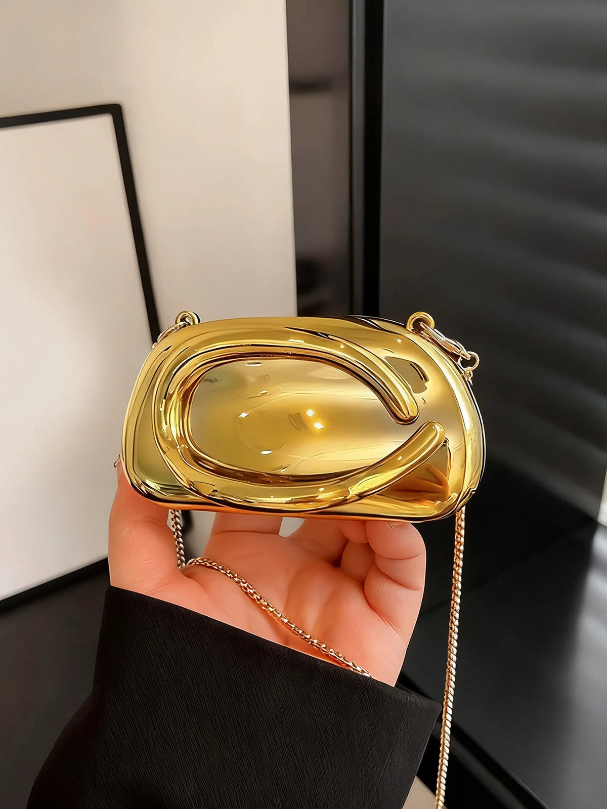 A hand holds a mini gold metal clutch bag with a gold chain strap against a dark background.