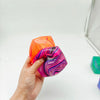 Une main tient un cube de jeu en gel anti-stress marbré orange et violet.