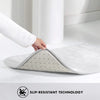 A white non-slip bathroom mat is shown on a white floor, with a hand placing it down.