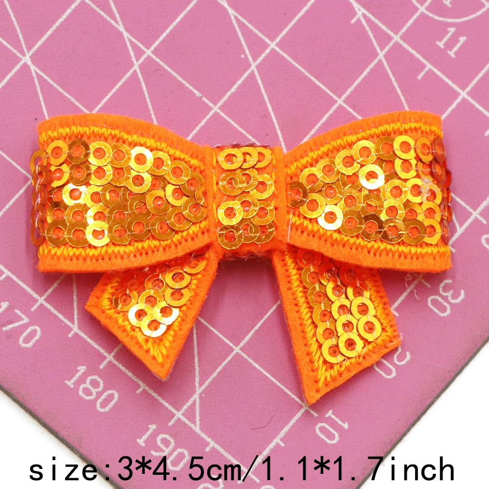A set of five orange sequin bow patches for DIY crafts displayed on a pink grid background.