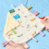 A hand holds a white baby sensory tag comfort blanket with various colored teethers on a light blue surface.