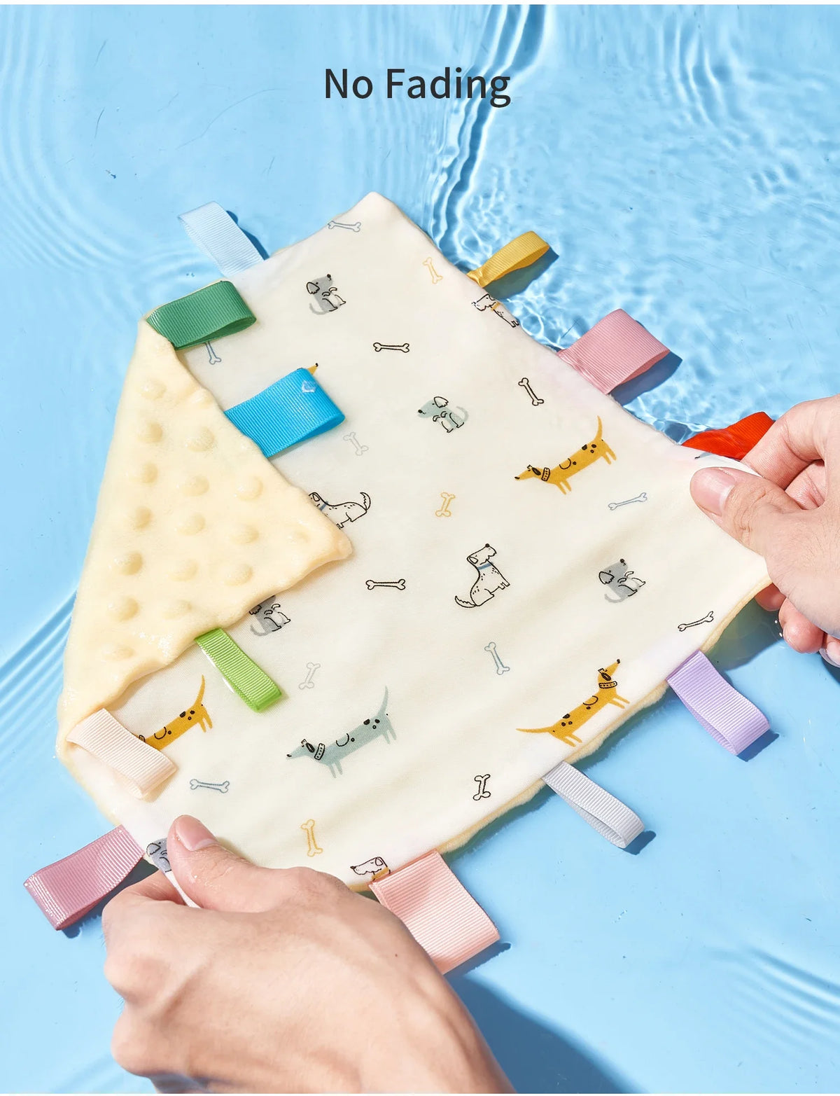 A hand holds a white baby sensory tag comfort blanket with various colored teethers on a light blue surface.