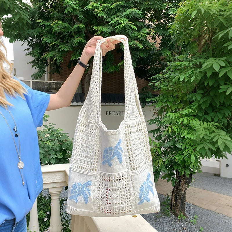 A person holds up a white handwoven knit tote bag with a hollow design outdoors.