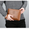Eine Person hält eine braune Kunstleder-Herren-Clutch-Geldbörse vor einem grauen Pullover und schwarzen Hosen.
