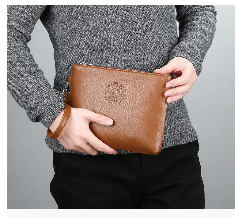 Eine Person hält eine braune Kunstleder-Herren-Clutch-Geldbörse vor einem grauen Pullover und schwarzen Hosen.