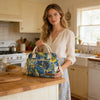 A woman holds a blue and yellow insulated picnic lunch bag with a light strap in a bright kitchen setting.