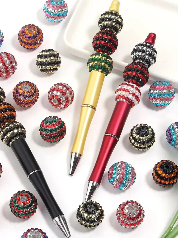 A set of five resin rhinestone beads with a two-tone striped design are displayed alongside a gold and red pen.