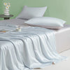 A light blue satin quilt draped over a bed with white pillows and a pale green wall background.