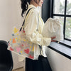 A person holds a white canvas tote bag and a matching transparent tote bag with an inner pouch next to a window.