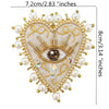 A heart-shaped patch featuring a gold-toned eye design surrounded by white pearls and clear rhinestones on a beige fabric.