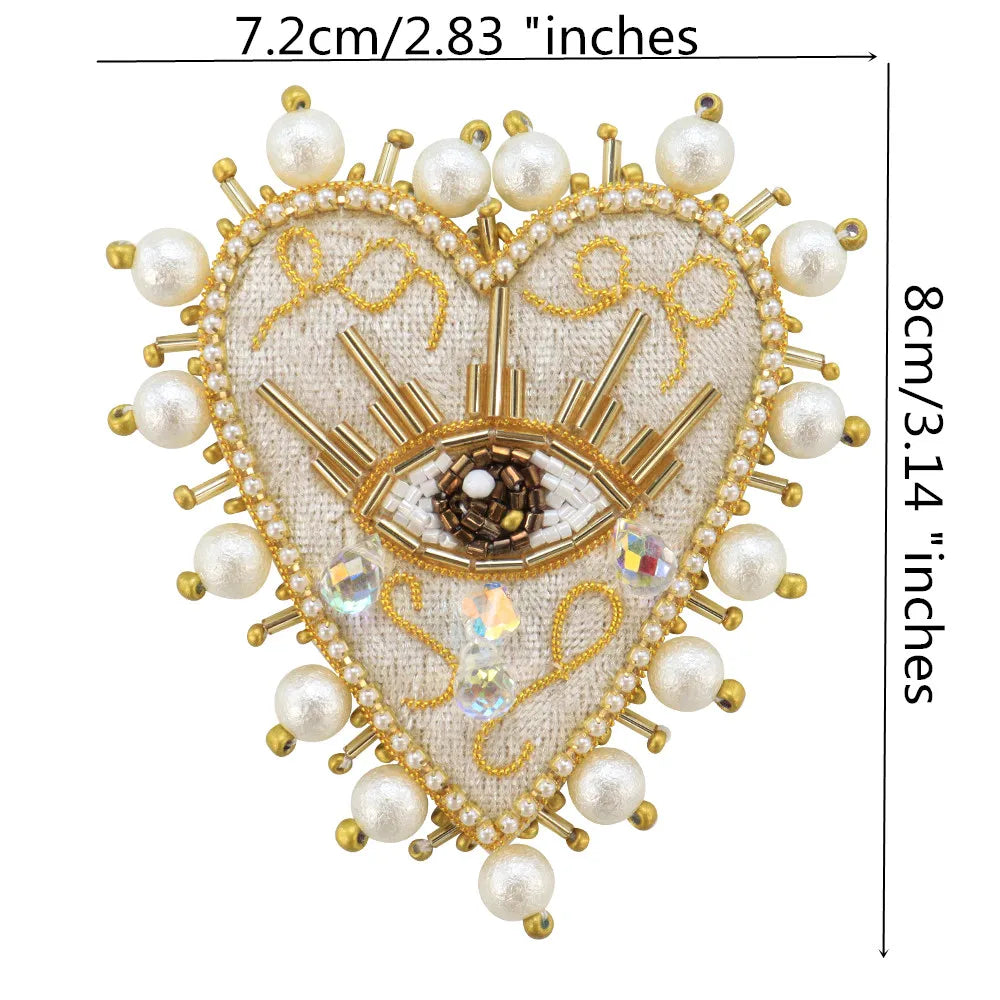 A heart-shaped patch featuring a gold-toned eye design surrounded by white pearls and clear rhinestones on a beige fabric.