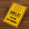 Un letrero amarillo con una cara sonriente y el texto 'Smile You're on camera' sobre una superficie de madera.