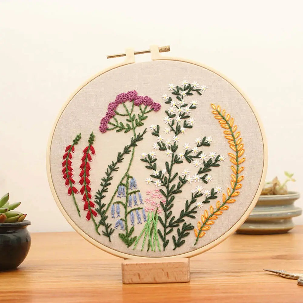 A circular embroidery hoop featuring floral cross stitch designs in shades of green, pink, white, and yellow on a beige fabri