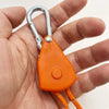 A hand holds an orange camping rope tensioner with a silver pulley buckle attached.