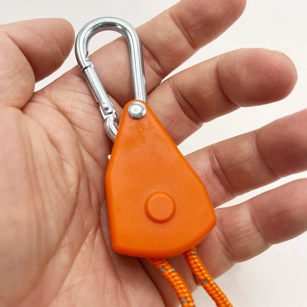A hand holds an orange camping rope tensioner with a silver pulley buckle attached.