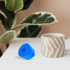 A blue silicone water jug cap resting on a white surface next to a beige and white striped mug.
