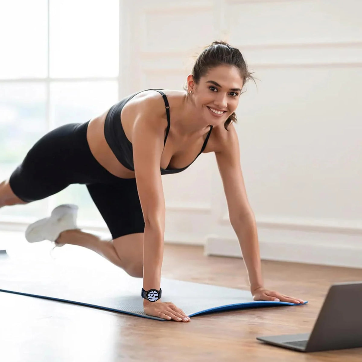 Una mujer con ropa deportiva negra realiza un estiramiento o ejercicio en una esterilla de yoga en interiores.