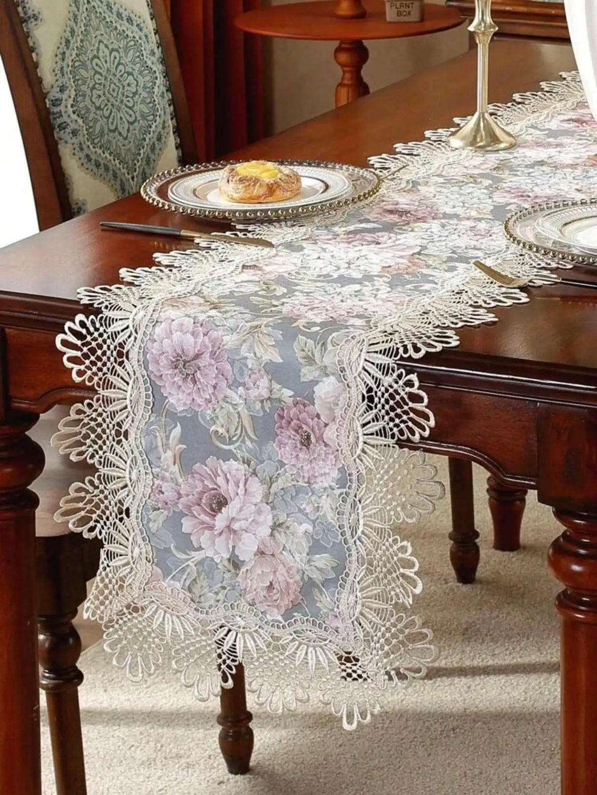 A lace table runner featuring a floral pattern in pink and gray on a dark wood table.