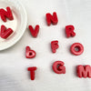 Perles de lettres en acrylique rouges formant N V R F O G T GM, éparpillées sur une surface blanche