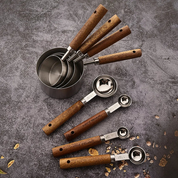 A set of stainless steel measuring cups and spoons with wooden handles arranged on a dark gray surface.