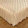 A beige quilted bed skirt with a pleated bottom and gold embroidery lies on a wooden floor.