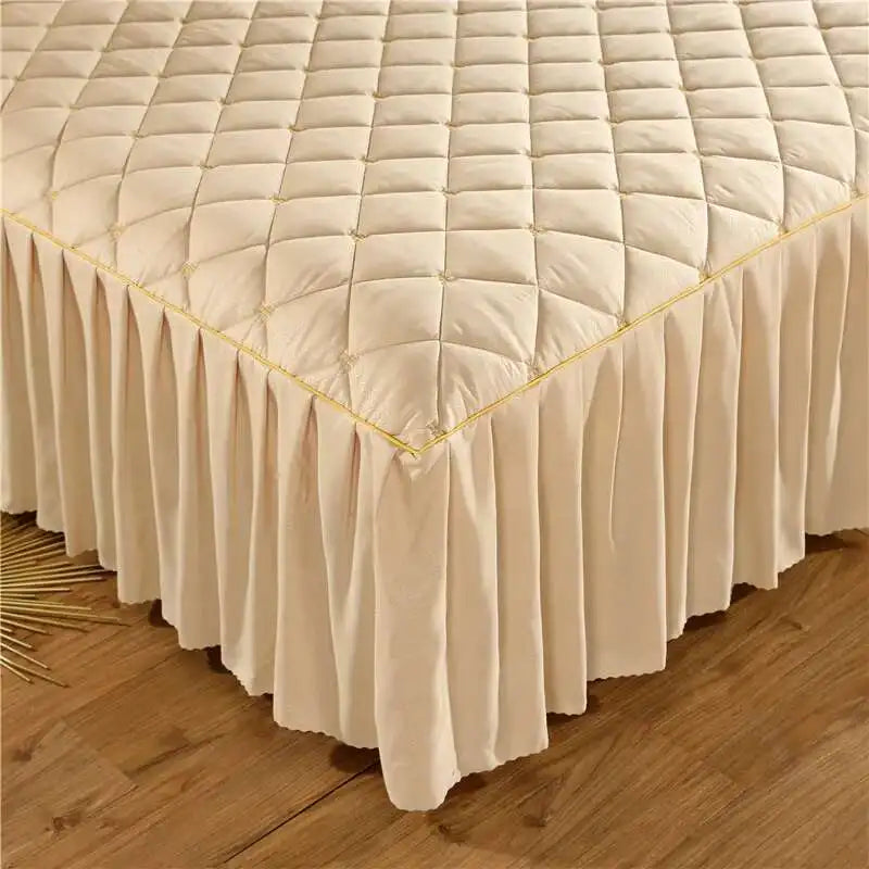 A beige quilted bed skirt with a pleated bottom and gold embroidery lies on a wooden floor.