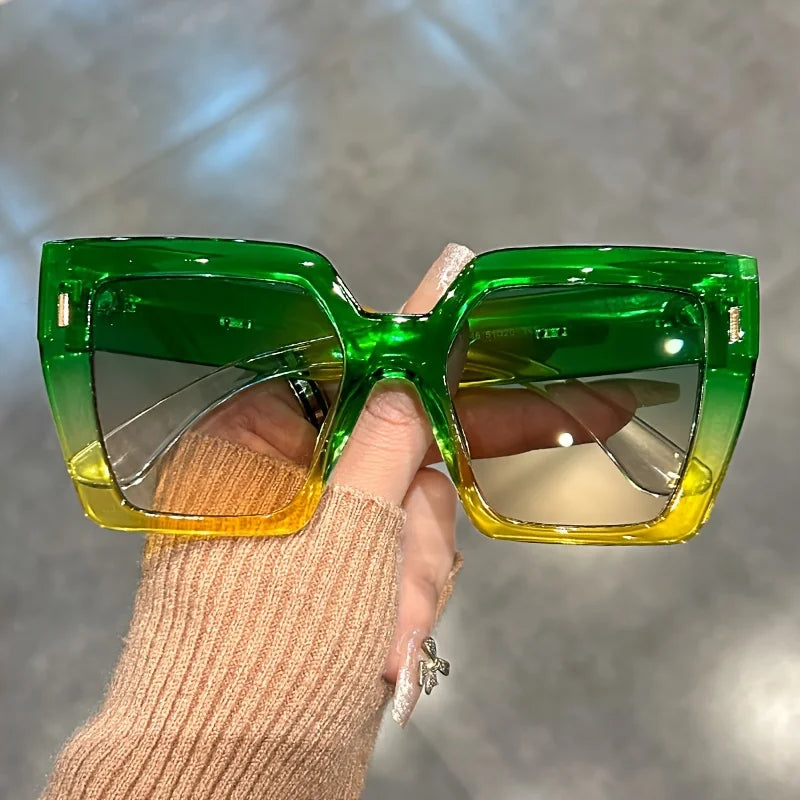 A hand holds a pair of bright green gradient square sunglasses against a blurred gray background.