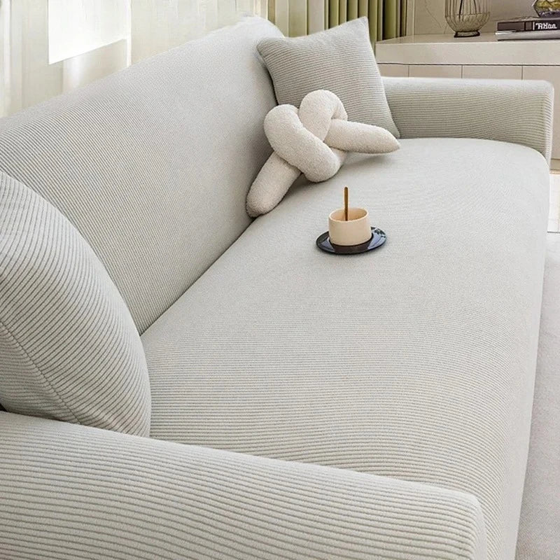 A light beige stretch jacquard sofa cover with a textured weave and a decorative knitted pillow on a sofa.