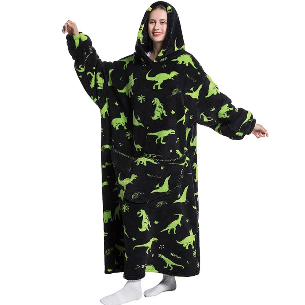 A woman wearing an oversized black hoodie blanket featuring a pattern of green dinosaurs and tropical leaves.