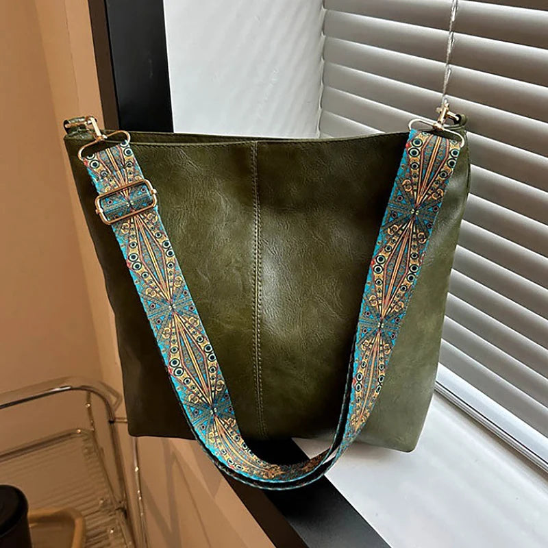 Sac à l'épaule vert olive foncé avec une sangle géométrique large et broderies multicolores devant une fenêtre.
