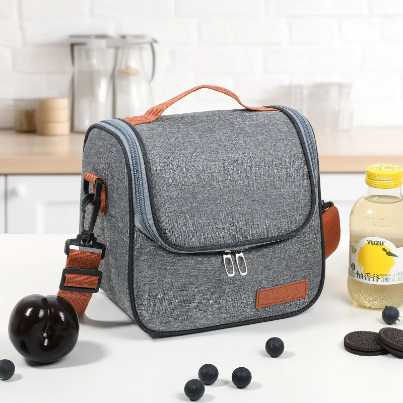 A gray insulated lunch bag with an orange strap is shown on a white surface next to several black fruit and cookie items.