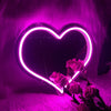 A glowing neon heart sign illuminates pink feathers and roses in a dark room.