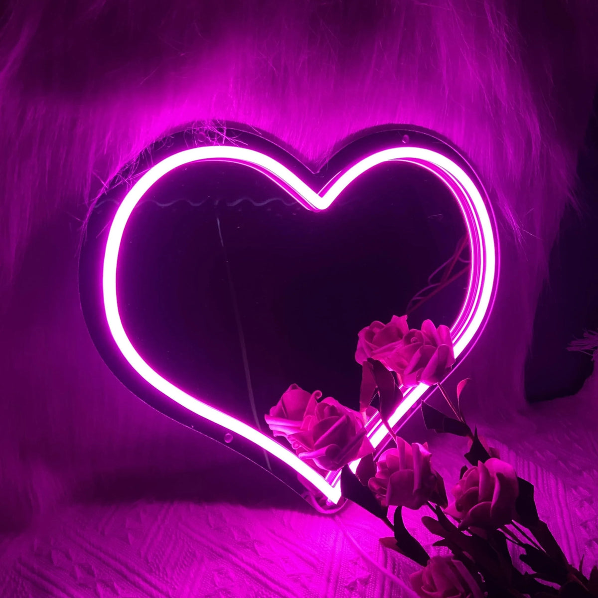 A glowing neon heart sign illuminates pink feathers and roses in a dark room.