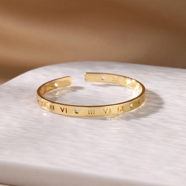 Bracelet manchette en acier inoxydable ton or gravé de chiffres romains sur une surface blanche.