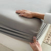 A hand is adjusting a gray cotton fitted sheet with an elastic band on a bed.