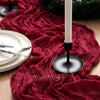 A deep red crinkle gauze table runner draped across a wooden surface with a white candle and greenery.