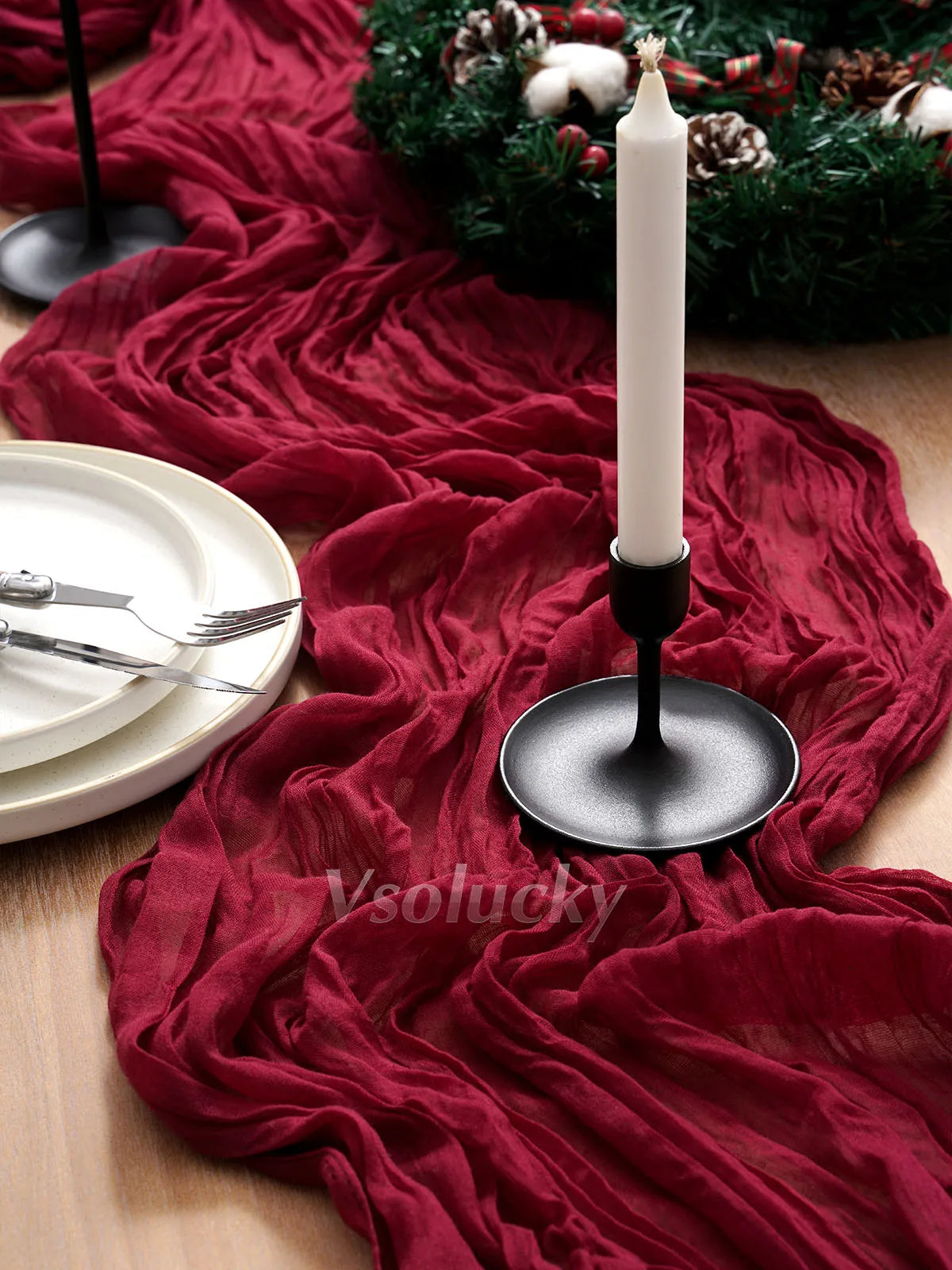 A deep red crinkle gauze table runner draped across a wooden surface with a white candle and greenery.