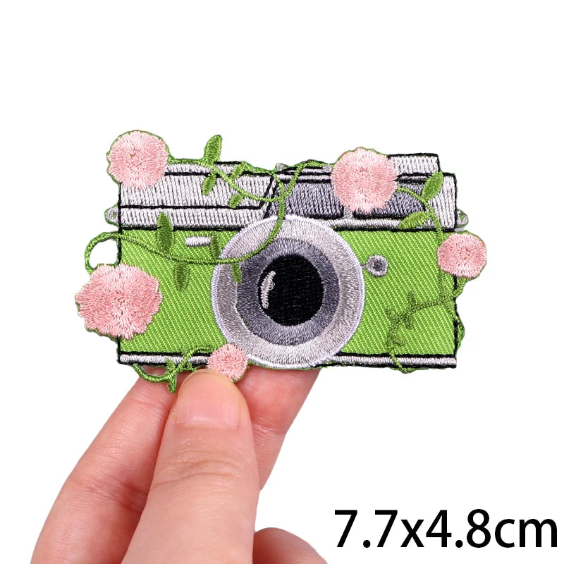 A hand holds a green embroidery patch featuring a cat face design with pink floral elements.