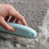 A hand is using a light blue pet hair removal brush on a gray, fuzzy surface.