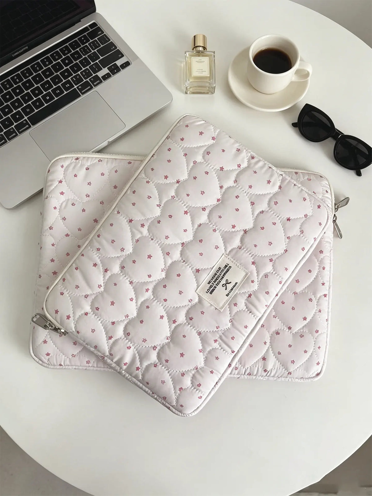 A quilted pink laptop sleeve with a zipper is placed next to a silver perfume bottle and a white cup on a light surface.