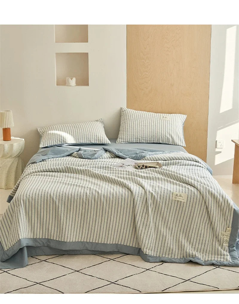 A light blue and white striped breathable summer quilt is displayed on a bed with a wooden headboard.