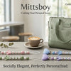 A green baby pacifier clip with a custom name is displayed on a wooden table next to various natural greenery.