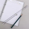 A transparent plastic ruler is placed diagonally across a spiral notebook with a grid, next to a dark pencil.