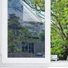 A close-up view of a silver one-way mirror window film installed in a white frame, showing greenery outside.