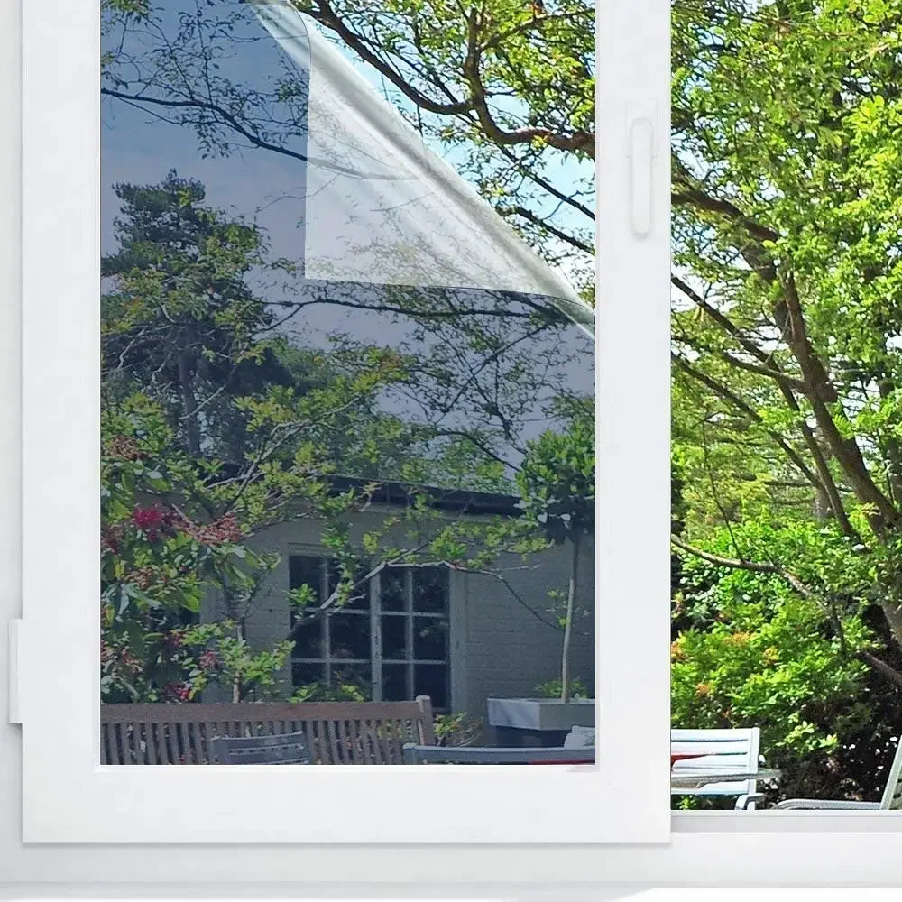 A close-up view of a silver one-way mirror window film installed in a white frame, showing greenery outside.