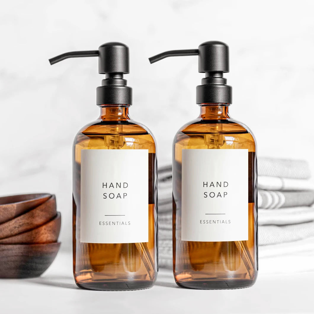 Two amber glass hand soap dispensers with black stainless steel pumps and labels on a white background.