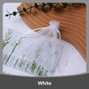 A white organza drawstring gift bag filled with small green plant sprigs, resting on a wooden surface.