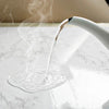 A white liquid is being poured from a white bottle onto a marble surface, creating a visible steam or mist.