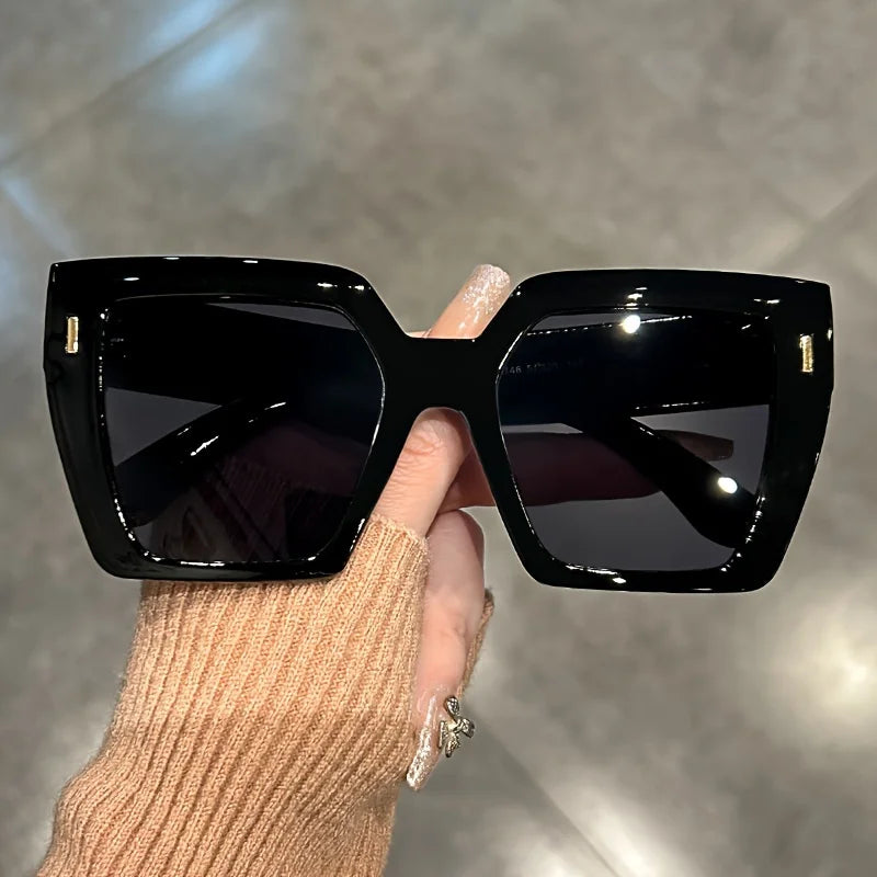 A hand holds a pair of black gradient square sunglasses against a blurred gray background.